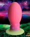 A whimsical, bright pink Xeno Egg Glow In The Dark Extra Large Silicone Egg stands on a green surface against a vibrant cosmic backdrop with planets and nebulae, giving off a surreal and fantastical vibe. (Brand: XR Brands)