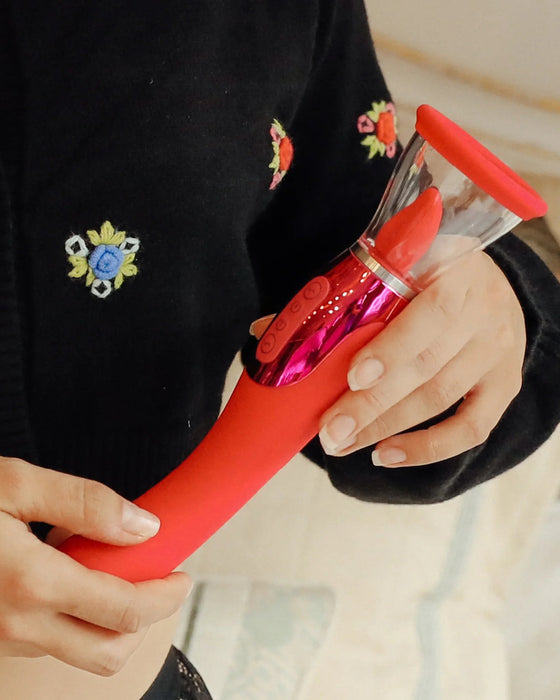 A close-up of a person's hands holding a pink Pipedream Products Jimmyjane Apex Double Ended Licking, Sucking G-Spot Vibrator with powerful motors and a clear container. The person is wearing a black sweater with colorful floral embroidery.