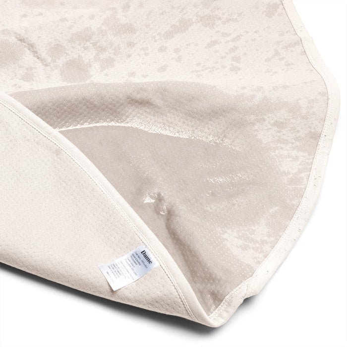 A close-up of the Dame Thro Soft Waterproof Sex Blanket, showing its light color, ultra-soft cotton top with a wet stain, flipped corner revealing the waterproof underside and fabric tag. The blanket is machine washable for easy care.