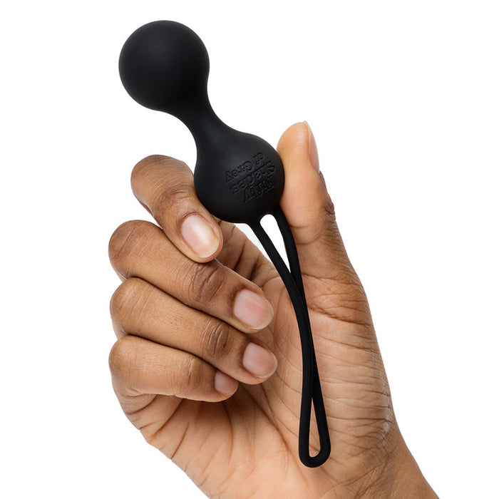 A hand holds a black silicone kegel exercise ball with a retrieval loop from the Fifty Shades of Grey Desire Blooms Kit, against a plain white background.
