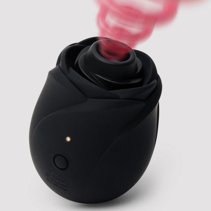 The Fifty Shades of Grey Hearts and Flowers Suction Rose is a black, rose-shaped clitoral stimulator with a glowing button, emitting pink swirling vapor upward. It uses air pulse technology for pleasure against a plain light background.