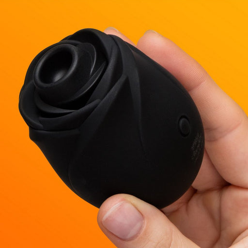 A hand holds the Fifty Shades of Grey Hearts and Flowers Suction Rose against an orange background, showcasing its elegant black design for clitoral stimulation.