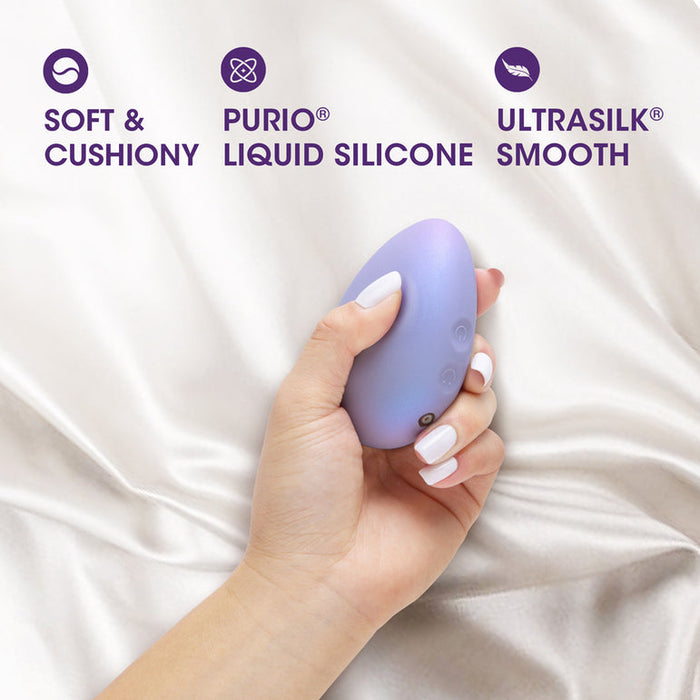 A hand with white-painted nails holds the Serene Silicone Waterproof Palm Vibrator, a small purple oval massager, against silky fabric. Text above reads: "Soft & Cushiony," "Purio Liquid Silicone," and "Ultrasilk Smooth.