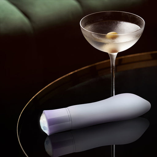 An Opal Heating Waterproof Silicone Vibrator in gray rests on a glossy black table beside a clear martini glass with an olive, with a dark green sofa in the background.