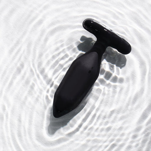 The Je Joue Onyx Large Powerful Remote Control Vibrating Butt Plug, in black silicone with a T-shaped base, rests on rippled white water, its circular patterns hinting at deep rumbly vibrations ready to be explored.