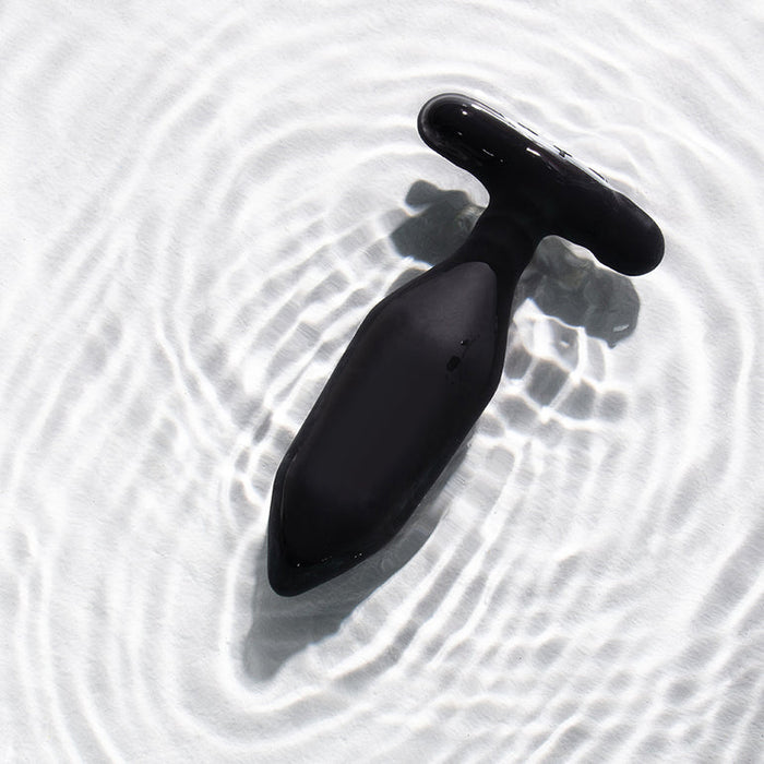 The Je Joue Onyx Large Powerful Remote Control Vibrating Butt Plug, in black silicone with a T-shaped base, rests on rippled white water, its circular patterns hinting at deep rumbly vibrations ready to be explored.