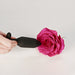 A hand holds the Je Joue Onyx Large Powerful Remote Control Vibrating Butt Plug near the center of a vibrant pink rose, both displayed against a plain white background.