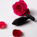 The Je Joue Onyx Large Powerful Remote Control Vibrating Butt Plug sits on a white surface beside a bright pink rose and petals, suggesting intense rumbly vibrations for an elevated sensual experience.