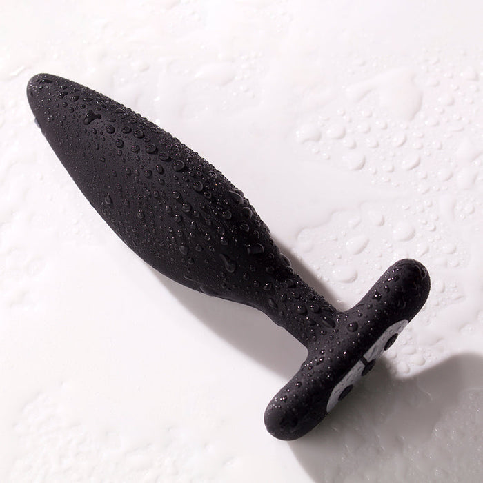 The Je Joue Egon Powerful Remote Control Vibrating Butt Plug, made of black silicone, glistens with water droplets on its surface as it rests on a wet white background.