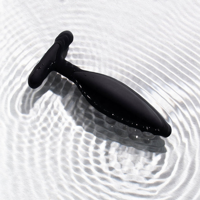 The Je Joue Egon Powerful Remote Control Vibrating Butt Plug, crafted from sleek black silicone with a handle, sits on a white surface as gentle circular ripples form in the water around it.