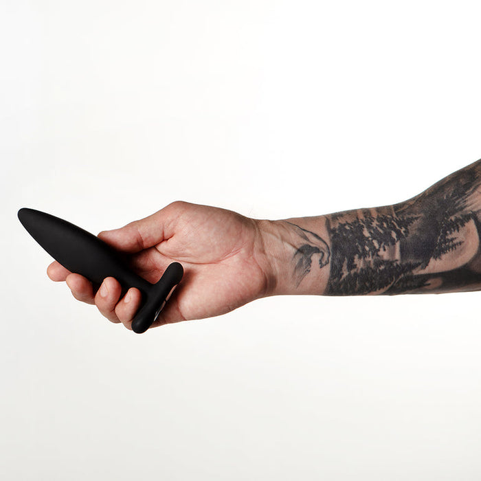 A tattooed arm holds the Je Joue Egon Powerful Remote Control Vibrating Butt Plug, made of black silicone, against a plain white background.