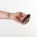 A hand with a mountain tattoo holds the Je Joue Vesta Petite Powerful Remote Control Vibrating Butt Plug's small, black remote against a plain white background. The plug is made from body-safe silicone and is oval-shaped.