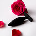 The Je Joue Egon Powerful Remote Control Vibrating Butt Plug in black silicone sits on a white surface, surrounded by red rose petals and a full red rose in the background.