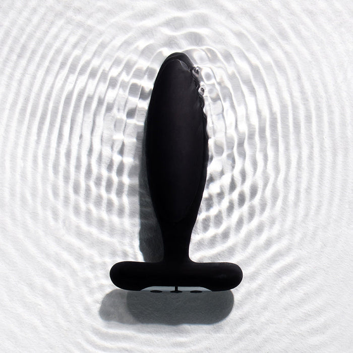 The Je Joue Vesta Petite Powerful Remote Control Vibrating Butt Plug—a black, body-safe silicone, T-shaped toy—stands upright on a textured white surface, creating ripples that suggest its waterproof design.
