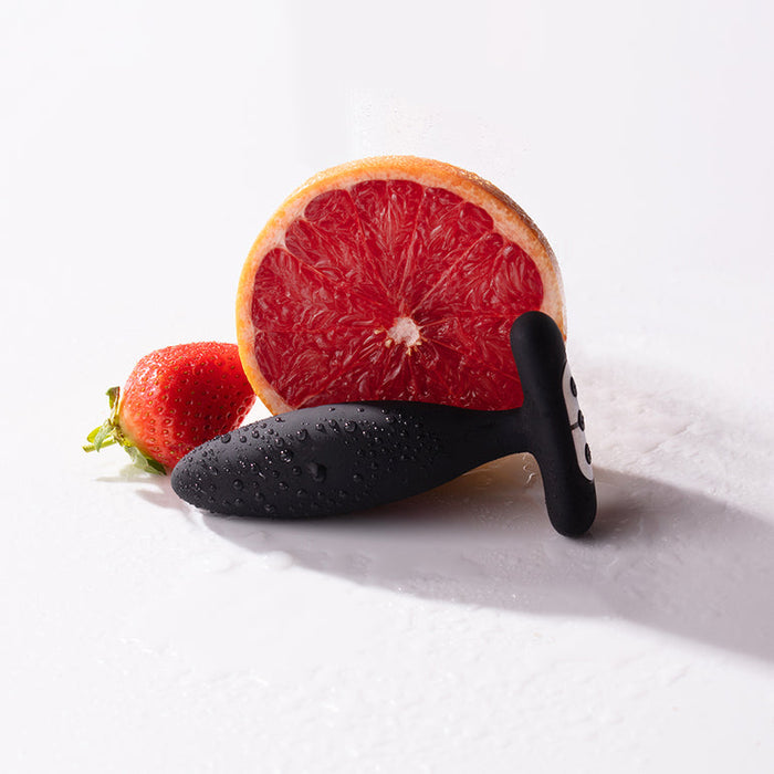 The Je Joue Vesta Petite Powerful Remote Control Vibrating Butt Plug, made of body-safe black silicone, sits with water droplets in front of a halved grapefruit and a whole strawberry on a white surface.