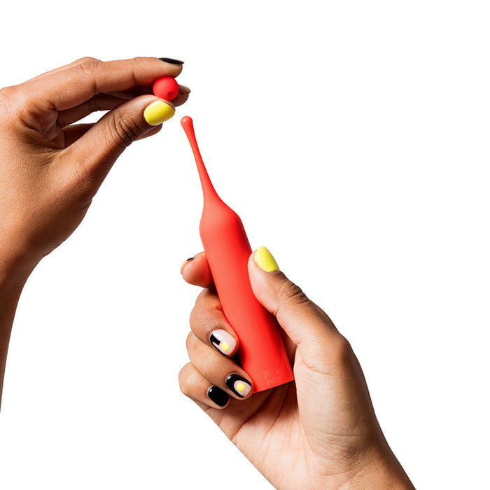 A person with yellow and black painted nails holds a red Romp Pop Pinpoint Clitoral Stimulator with 2 heads in one hand and a small red ball in the other, against a white background.