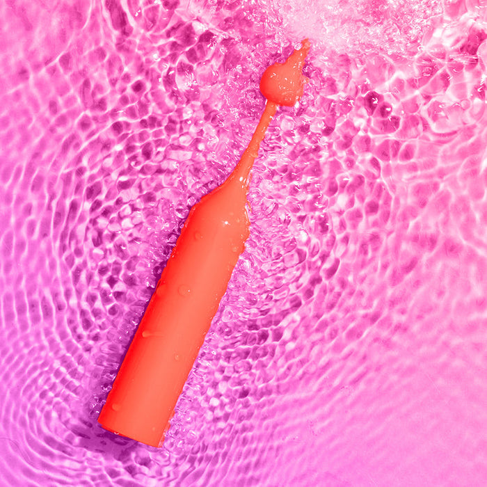The Romp Pop Pinpoint Clitoral Stimulator with 2 Heads, in coral, features water droplets on its surface as it rests on rippling water reflecting pink light.
