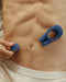 A close-up of a person's toned abdomen with a hand holding a Pipedream Products Jimmyjane Deimos Dual Motor Vibrating Cock Ring for Couples placed near the belly button. The person is wearing white undergarments.