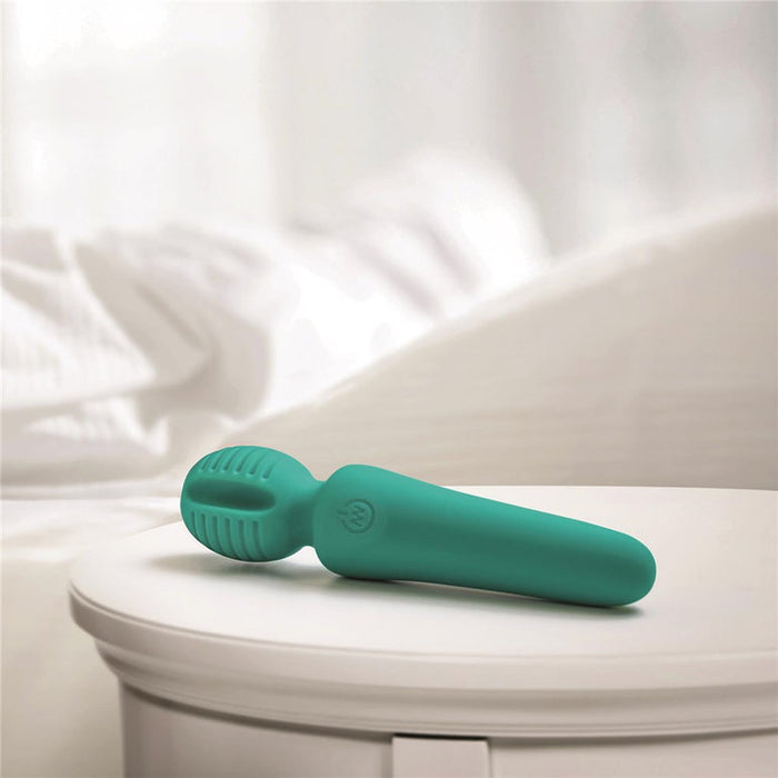 The Petite Private Pleasure Waterproof Silicone Wand Vibrator, in teal silicone, rests on a white round bedside table with a softly blurred bed and white bedding in the background.