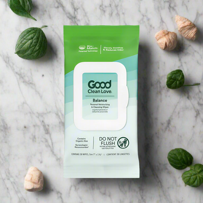 A 30-count pack of Good Clean Love Balance Personal Moisturizing & Cleansing Wipes is shown on marble with green leaves and dried beige flowers. The pH balanced packaging features aloe, freshness, and a "Do Not Flush" label.