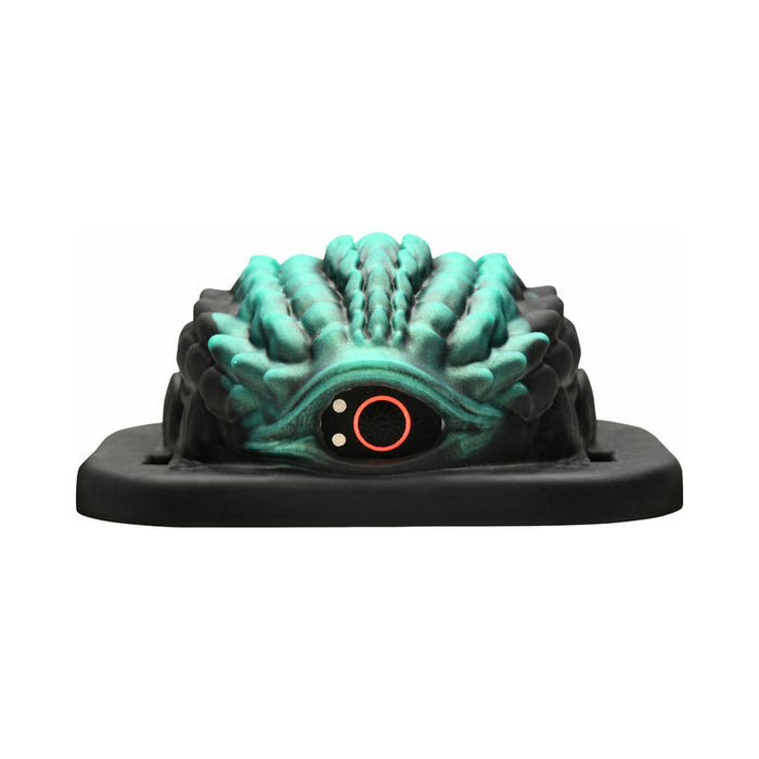 A cake modeled after the Dragon Grinder - Vibrating Silicone Fantasy Pad, with teal-green and black textured ridges, a prominent orange and black eye in front, sits on a black rectangular base.