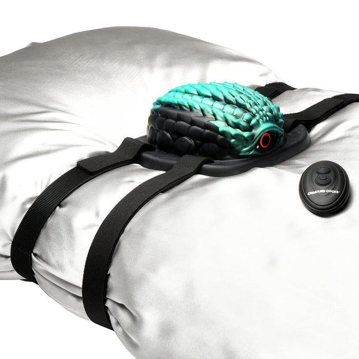 A gray pillow with black straps holds the turquoise and black Dragon Grinder - Vibrating Silicone Fantasy Pad, featuring dragon scale designs. Includes a small "Creature Couch" remote. This waterproof, rechargeable pad brings fantasy to life.