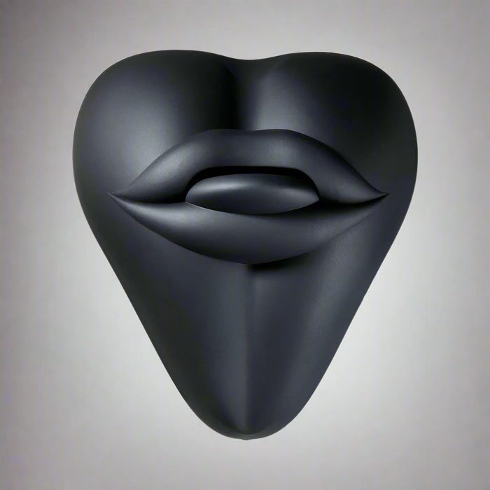 The Bananapants Kissi Dildo Base features a black, sculptural heart shape with abstract lips and tongue on its surface. Crafted from body-safe silicone, it's shown against a white background.