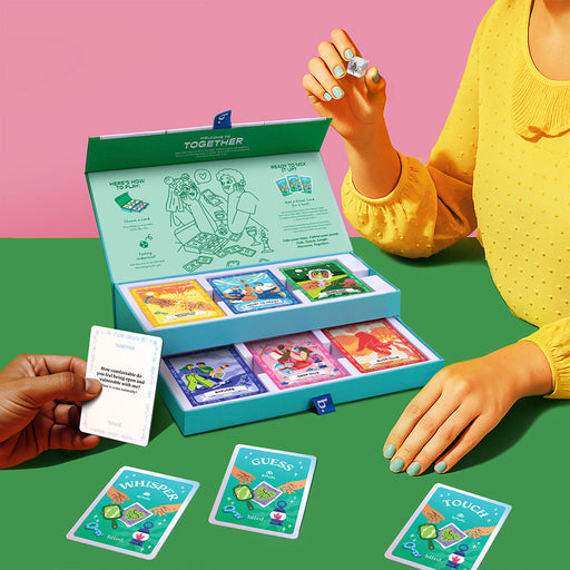 A person in a yellow top holds a die next to the open Together Couples Intimacy Card Game box with illustrated cards and dice. Another hand has a white card. Cards labeled "Whisper," "Guess," and "Touch" inspire communication for couples.
