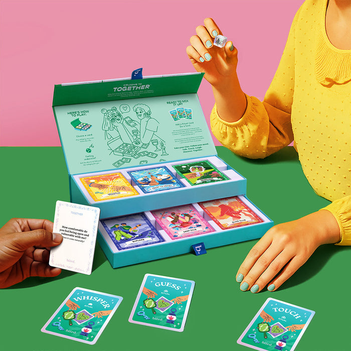 A person in a yellow top holds a die next to the open Together Couples Intimacy Card Game box with illustrated cards and dice. Another hand has a white card. Cards labeled "Whisper," "Guess," and "Touch" inspire communication for couples.