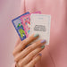 A hand with pastel green and lavender nails holds four cards from the Together Couples Intimacy Card Game; the front card reads, "How would you describe your love language, and has it changed over time?" on a soft pink fabric background.