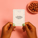 Two hands with mint green nail polish hold a Together Couples Intimacy Card Game card that says, "How do you define vulnerability, and how do you show it?" A pink bowl of pretzels sits on a pink surface in the background.
