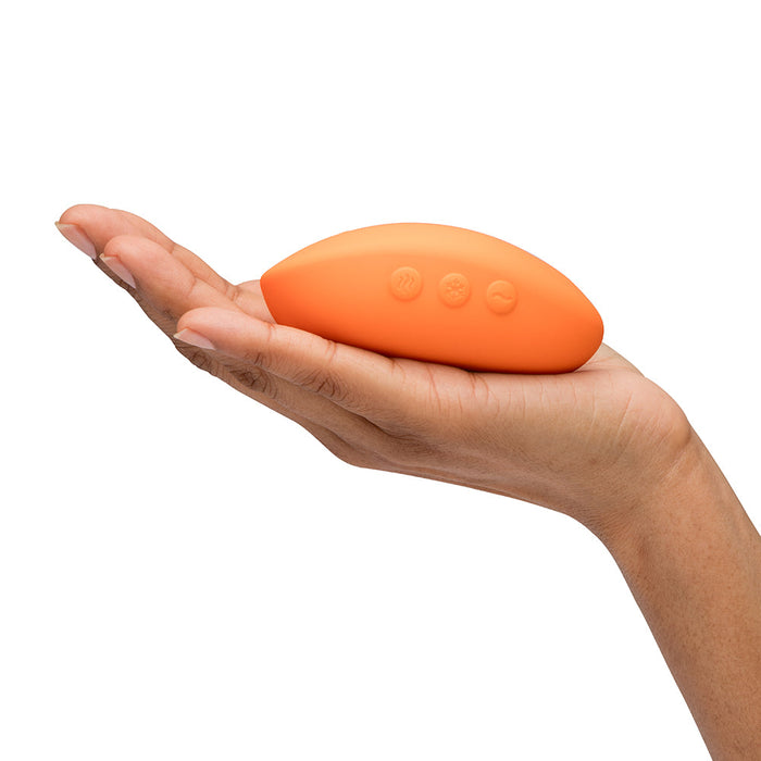 A hand holds the We-Vibe Temp Lay On Vibrator—an orange, oval device with three side buttons for hot and cold functions—against a white background.