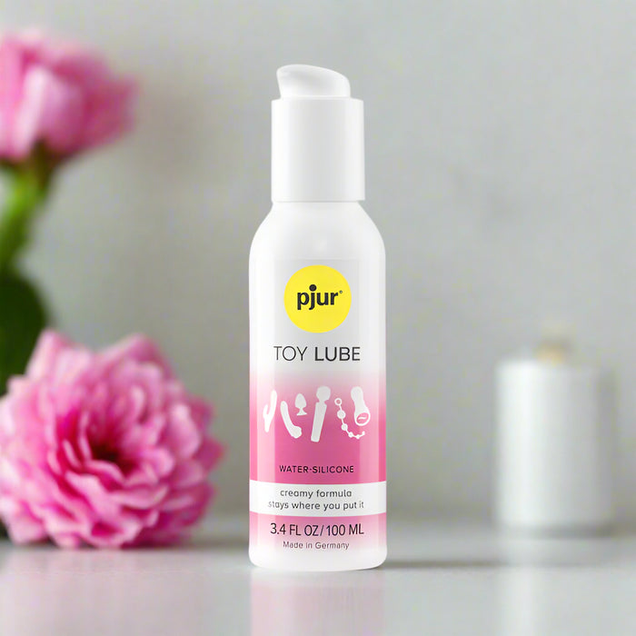 A bottle of Pjur Toy Lube Ultra Thick Silicone/Water Based Hybrid Lubricant for Sex Toys with a white and pink label sits on a reflective surface, while a pink flower and white candle blur in the background, emphasizing its long-lasting lubrication.