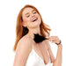 A smiling red-haired woman in a white satin top gently brushes her neck with the Sex & Mischief Black Wrist Feather Tickler, looking relaxed and joyful as she enjoys sensory play against a plain white background.