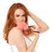 A woman with long red hair smiles at the camera, holding the Peaches 'n Cream Wrist Feather Tickler near her face while wearing a white satin camisole, inviting playful sensory exploration against a white background.