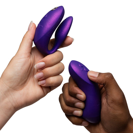 Two hands—one light-skinned, one dark-skinned—hold the We-Vibe Chorus Pro Wearable Couples' Vibrator with Triple Motors & Remote in purple against a white background. It features Fusion Wave Technology for precise G-spot stimulation and enhanced pleasure.
