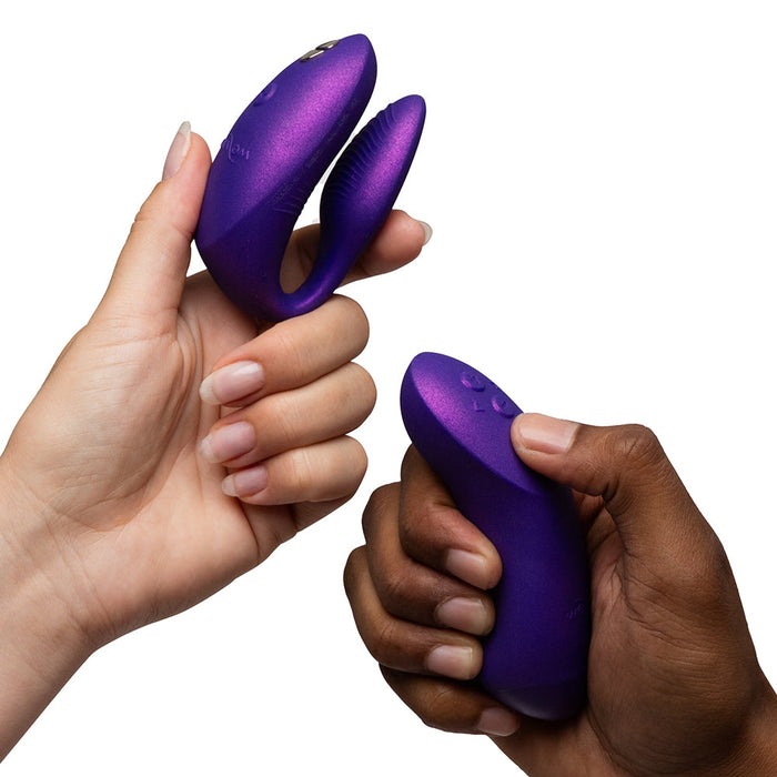 Two hands—one light-skinned, one dark-skinned—hold the We-Vibe Chorus Pro Wearable Couples' Vibrator with Triple Motors & Remote in purple against a white background. It features Fusion Wave Technology for precise G-spot stimulation and enhanced pleasure.
