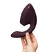 A hand holds the Womanizer Next Duo Pleasure Air + Vibration Blended Orgasm Stimulator, a dark purple, curved personal massager, against a plain white background.
