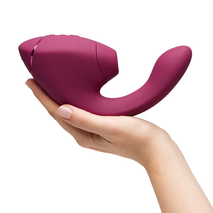 A hand holds the maroon Womanizer Next Duo Pleasure Air + Vibration Blended Orgasm Stimulator against a plain white background.