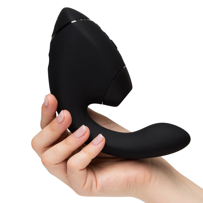 A hand holds the Womanizer Next Duo Pleasure Air + Vibration Blended Orgasm Stimulator, a black, curved silicone device with an ergonomic contoured handle and bulbous end, shown against a plain white background.