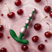 The Bougie Vibrating Aluminum Anal Beads in green and pink rest on a glossy pink surface, surrounded by fresh cherries and cherry juice.