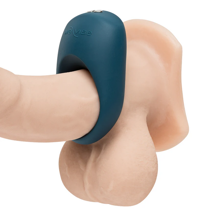 A We-Vibe Pivot 2 Vibrating Silicone Rechargeable Penis Ring in blue is worn at the base of a realistic, flesh-toned dildo and testicles, delivering hands-free clitoral stimulation against a white background.