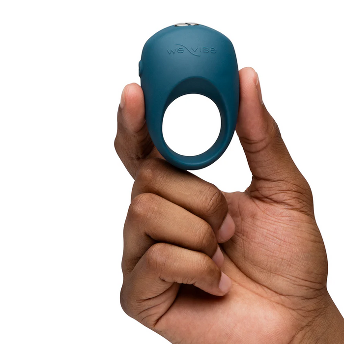 A hand holds up the We-Vibe Pivot 2 Vibrating Silicone Rechargeable Penis Ring, a blue ring designed for hands-free clitoral stimulation, with the "We-Vibe" logo visible against a white background.