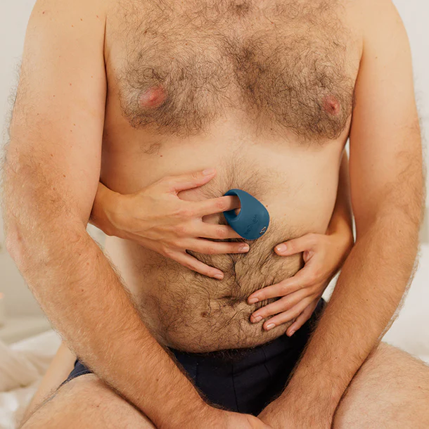 A shirtless man in underwear sits with another person’s hands on his stomach, one hand pressing the We-Vibe Pivot 2 Vibrating Silicone Rechargeable Penis Ring to his lower abdomen for shared pleasure.