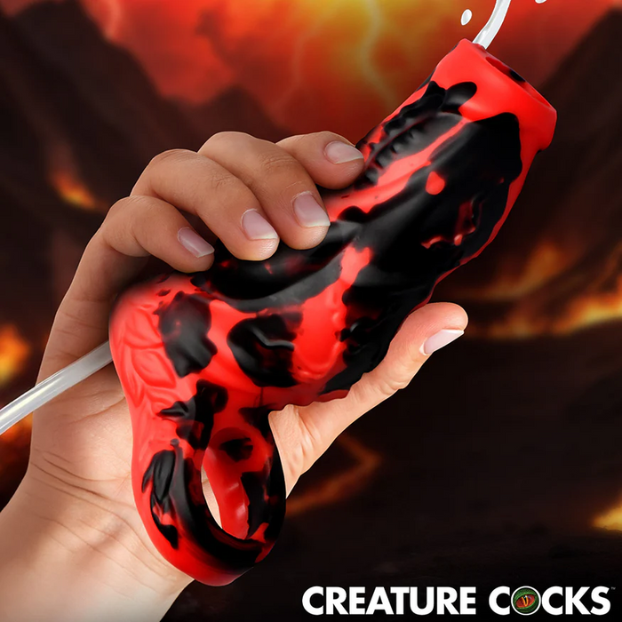 A hand holds the Hell Hound Canine Squirting Penis Enhancer, a red and black fantasy toy with an attached tube, against a blurred fiery mountainous background. "CREATURE COCKS" appears at the bottom of the image.