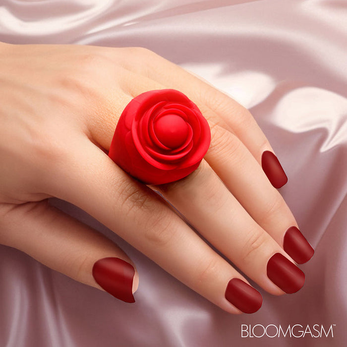 A hand with matte red nails wears the Bloomgasm Rose Ring Finger Vibrator, shaped like a blooming rose from body-safe silicone, on smooth pale pink satin. "BLOOMGASM" is displayed in the bottom right corner.