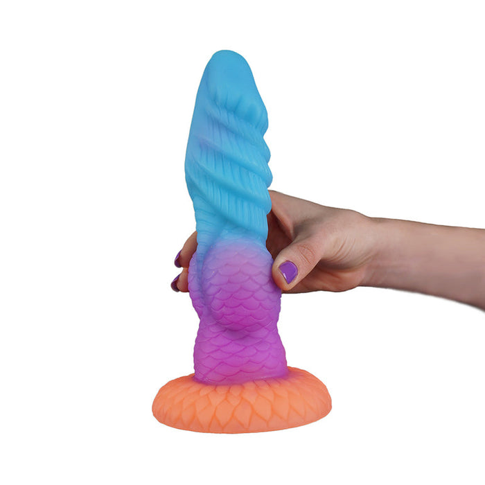 A hand with purple-painted nails holds the Splash Nova Squirting 10 Inch Glow in the Dark Creature Dildo, featuring a colorful, textured, scale-like design and a wide orange base, set against a white background.