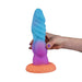 A hand with purple-painted nails holds the Splash Nova Squirting 10 Inch Glow in the Dark Creature Dildo, featuring a colorful, textured, scale-like design and a wide orange base, set against a white background.