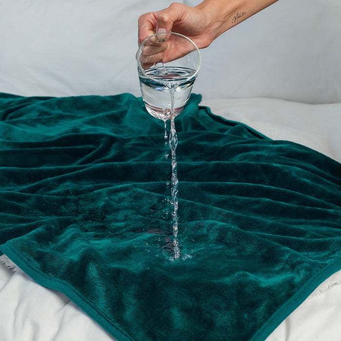 A hand pours water onto the Splash Blanket The OG Waterproof Blanket - Small in dark green, demonstrating its effective spill protection and impressive absorbency on a bed.