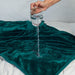 A hand pours water onto the Splash Blanket The OG Waterproof Blanket - Small in dark green, demonstrating its effective spill protection and impressive absorbency on a bed.